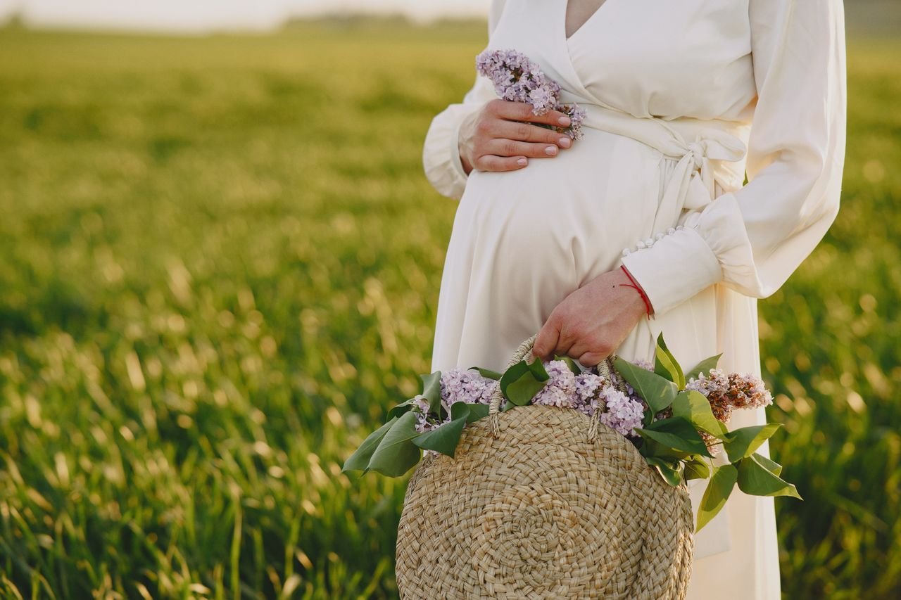 A pregnant woman in a white dress holds a straw bag with flowers in a lush green field.